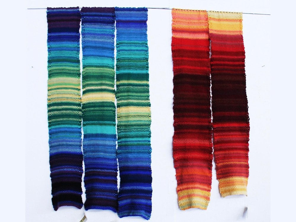 Five narrow textiles are hung vertically. They are striped and have a variety of colors: Blue, green, yellow, red and orange.