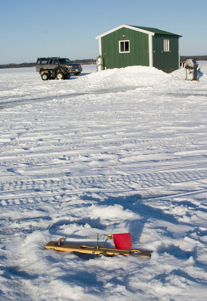 Photo of a frozen lake with a green ice fishing shed. Snow in the foreground had tire tracks and two vehicles are parked near the shed.